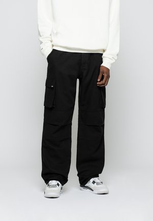 Czarne spodnie bojówki z dużymi bocznymi kieszeniami oraz białe trampki na osobie ubranej w białą bluzę, stojącej z rękami w kieszeniach.