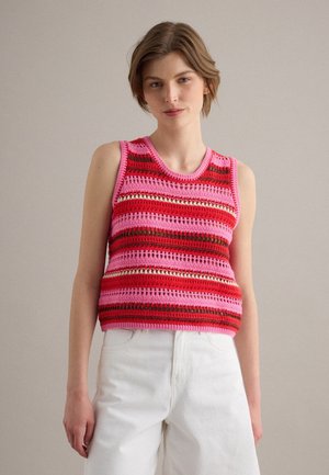 Giovane donna con top lavorato a uncinetto senza maniche a righe rosa, rosse e marroni abbinato a pantaloni bianchi a vita alta, in piedi davanti a uno sfondo neutro.