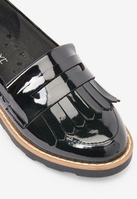 Zwarte lakleren loafers met franjes, ronde neus en contrasterende beige stiksels op een dikke zwarte zool.