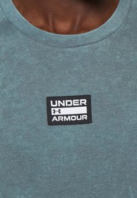 Tyrkysové bavlněné tričko s kulatým výstřihem a černou etiketou s bílým textem "UNDER ARMOUR" vyšitou na přední straně.