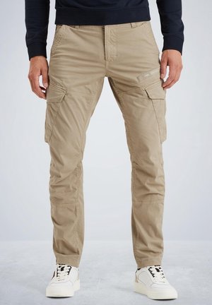 Hombre vistiendo pantalones cargo beige con múltiples bolsillos y zapatillas blancas, de pie contra un fondo claro y liso.