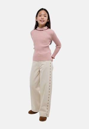 Jeune fille debout avec les mains derrière le dos, portant un pull côtelé rose, un pantalon large beige avec des détails floraux sur les côtés, et des chaussures marron.