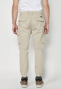 Beige Cargo-Hosen aus Baumwolle mit elastischem Knöchelbund, ausgestattet mit Seitentaschen und einer aufgesetzten Gesäßtasche.