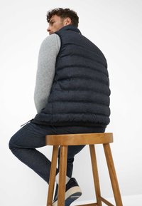 Navy Puffer-Weste mit horizontaler Steppung, hohem Kragen und schwarzem Saum. Über einem grauen Sweater getragen, kombiniert mit strukturierten navyfarbenen Hosen und Schuhen.