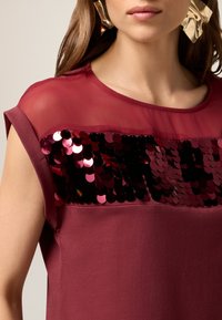 Haut bourgogne avec une superposition transparente et des embellissements en sequins sur la poitrine, manches courtes, texture douce et décolleté rond.