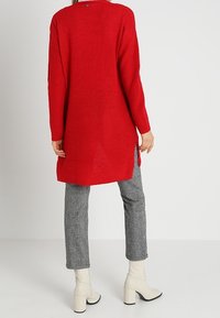Long pull rouge en maille avec une coupe décontractée, doté d'un col rond et de fentes latérales. Associé à un pantalon gris à carreaux et des bottines blanches.