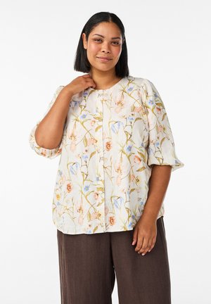 Žena so stredne dlhými čiernymi vlasmi, ktorá má na sebe krémovú kvetovanú blúzku a hnedé nohavice, pózuje s jednou rukou blízko krku.