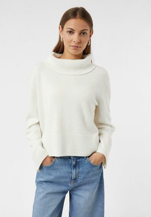 Maglione bianco a costine con ampio collo a caduta e maniche lunghe, abbinato a jeans blu. Il tessuto sembra morbido e leggermente testurizzato.