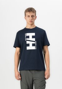 Junger Mann, der ein marineblaues Helly Hansen T-Shirt mit großem weißem Logo-Druck trägt, kombiniert mit grauen Cargo-Hosen, steht vor einem weißen Hintergrund.