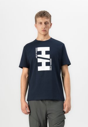 Junger Mann, der ein marineblaues Helly Hansen T-Shirt mit großem weißem Logo-Druck trägt, kombiniert mit grauen Cargo-Hosen, steht vor einem weißen Hintergrund.