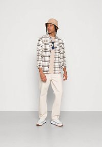 Un tânăr poartă o pălărie de tip bucket bej, o cămașă în carouri crem peste un tricou grafic bej, pantaloni crem și adidași albi, stând în fața unui perete simplu.