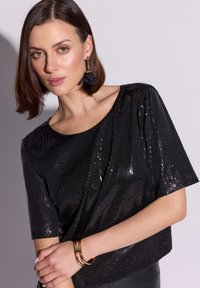 Czarny top z cekinami o luźnym kroju i krótkich rękawach, z okrągłym dekoltem. Uzupełniony złotą bransoletką i teksturowanymi kolczykami.