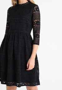 Svartrandad klänning i svart spets med trekvartsärmar, en figurnära överdel och en utställd kjol, med en scalloped ringning och intrikata mönster.