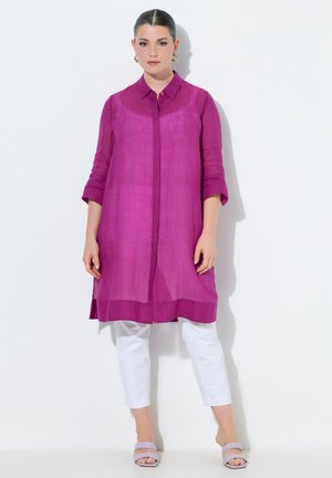 Nainen, joka pukeutunut pitkään, läpikuultavaan magentanväriseen tunikaan, jossa on käärityt hihat, valkoisiin polvipituisisiin housuihin ja laventelimaisiin avoimiin sandaaleihin, seisoo valkoisen seinän edessä.