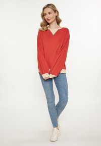 Punainen pitkähihainen paita, jossa on bégen värinen kaula-aukko ja hihansuut, yhdistettynä vaaleansinisiin slim fit -farkkuihin ja valkoisiin lenkkareihin. Yksinkertainen muotoilu, pehmeä rakenne.