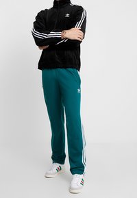 Schwarze Cordjacke mit Reißverschluss, weißen Streifen und Logo. Türkise Trainingshose mit weißen Streifen und seitlichen Taschen. Weiße Sneakers mit farbigen Akzenten.