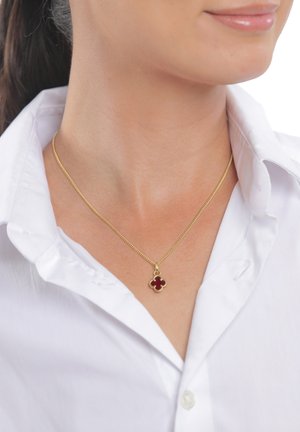 Femme portant un collier en or avec un pendentif trèfle à quatre feuilles rouge sur une chemise blanche à col, montrant le cou et les lèvres.
