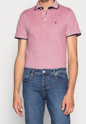 Homme portant un polo à manches courtes rose clair avec des bordures bleu marine et un jean en denim bleu, debout devant un fond uni clair.