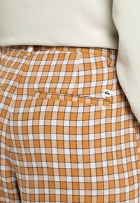 Bruine en witte geruite broek van een gewaxt stof. Heeft een achterzak en een klein logo-label op de tailleband.