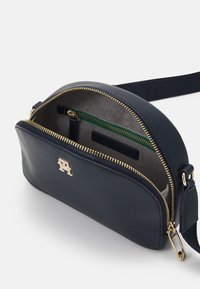 Borsa a tracolla in pelle blu navy con zip dorata, accento con logo dorato e una tasca interna con zip verde. Presenta una texture liscia e una forma arrotondata.