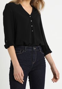 Kvindelig i en sort V-hals bluse med optrillede ærmer og mørkeblå højtaljede jeans, stående foran en ensfarvet baggrund.