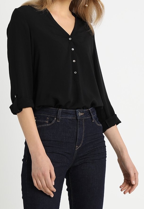 Kvindelig i en sort V-hals bluse med optrillede ærmer og mørkeblå højtaljede jeans, stående foran en ensfarvet baggrund.