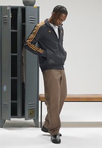 Σκούρα γκρι αθλητική ζακέτα με χρυσές ρίγες και λογότυπο, καφέ παντελόνια και μαύρα αθλητικά παπούτσια, μπροστά από μια μεταλλική ντουλάπα και πάγκο.