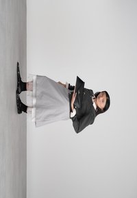 Noor naine hallis kleidis ja mustas jakis, hoiab musta kotti, jalas mustad saapad valgete pitsist sokkidega, seisab hallil põrandal.