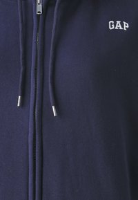 Sudadera con cremallera color azul marino hecha de suave tela de algodón, que presenta un pequeño logo blanco "GAP", cremallera plateada y cordones con puntas metálicas.