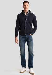 Cardigan lavorato a maglia blu con bottoni, cappuccio parziale e taschino sul petto, abbinato a jeans blu e sneakers bianche. Design pulito e vestibilità casual.