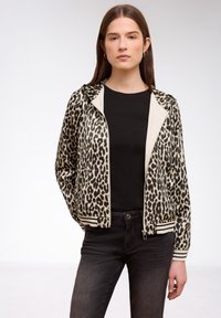 Junge Frau mit langen braunen Haaren, die Creolen trägt, eine Reißverschlussjacke mit Leopardenmuster über einem schwarzen Shirt und dunkle Jeans vor einem schlichten Hintergrund.