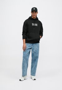 Hoodie noir avec une poche avant et un graphique blanc, associé à un jean bleu clair déchiré et des baskets blanches. Casquette noire avec logo.