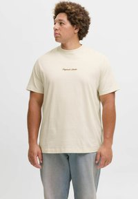 T-shirt en coton beige avec un col rond et des manches courtes. Présente l'inscription "Original Studio" brodée en marron sur la poitrine. Coupe décontractée.