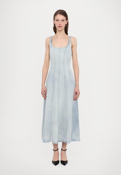 Robe en denim bleu clair avec un corsage ajusté et une jupe en forme A, présentant des bretelles étroites et des détails de coutures visibles. Chaussures noires à talons aiguille.