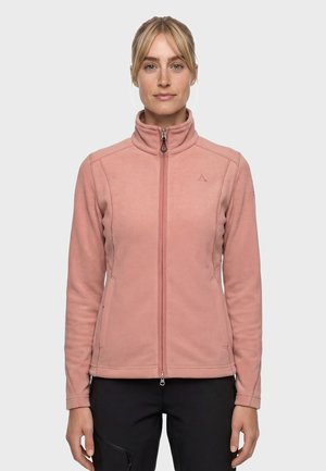 Frau trägt eine hellrosa Fleecejacke mit Reißverschluss und hohem Kragen sowie schwarze Hose, steht vor einem einfarbig hellen Hintergrund.