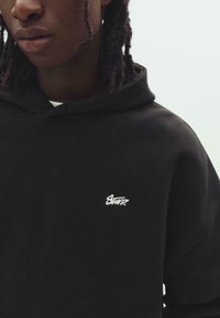 Jeune personne avec des cheveux tressés foncés portant un sweat à capuche noir avec un petit logo blanc "Stuntz" sur la poitrine, sur fond uni.