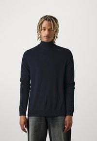 Pullover de gola alta em azul-marinho com mangas longas, punhos e bainha canelados, feito de um tecido suave, combinado com jeans cinza ligeiramente desgastados.