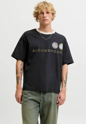 Personne blonde avec des tatouages porte un t-shirt noir avec le texte "ALCOBENDAS" et des badges, un pantalon vert, un collier chaîne et des boucles d'oreilles anneaux dorées.