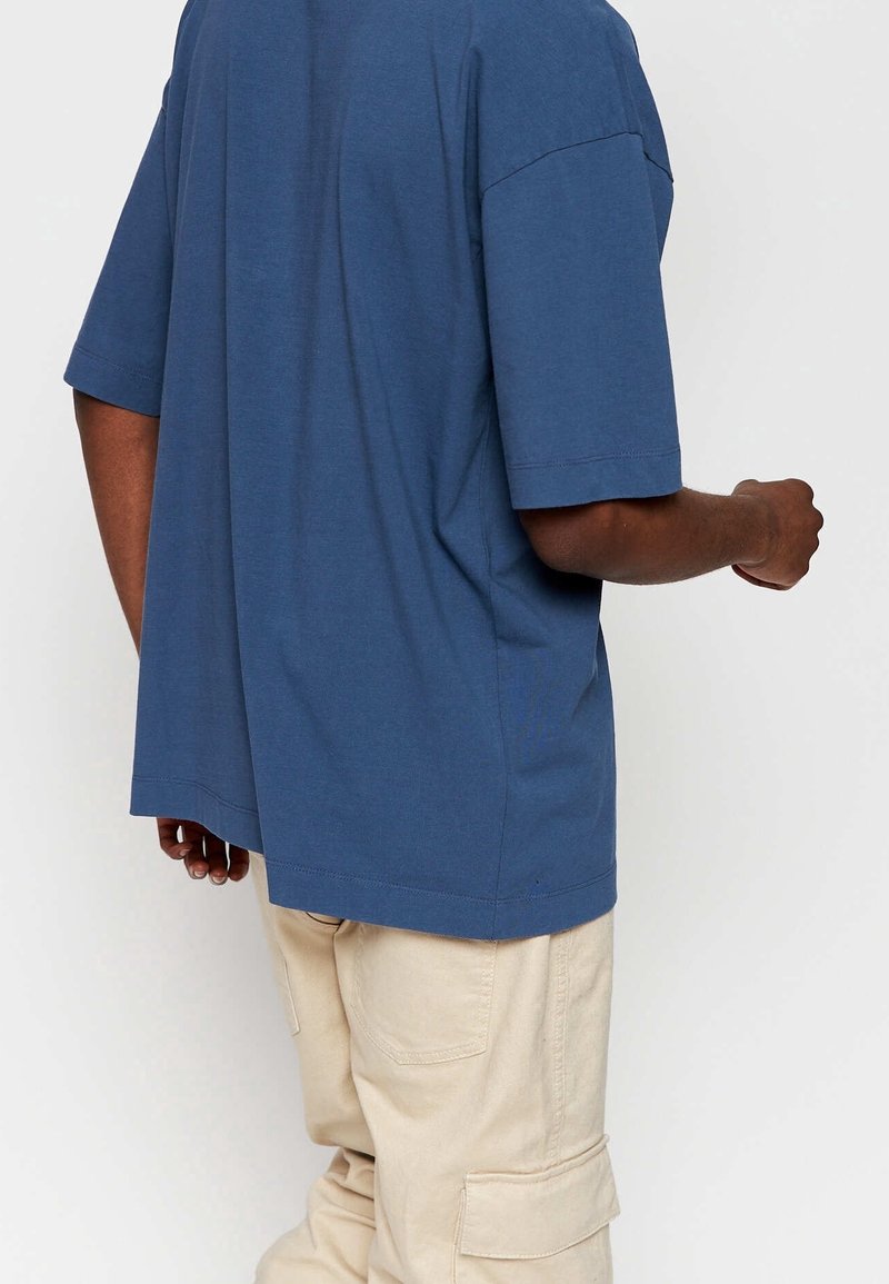 Personne portant une chemise bleu clair à manches courtes ample et un pantalon cargo beige clair, vue de dos sur un fond uni.