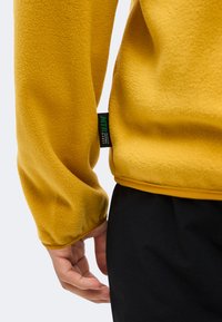 Κίτρινο μανίκι fleece μπουφάν με πράσινη και μαύρη ετικέτα εμπορικού σήματος, που βρίσκεται δίπλα σε ένα χέρι και μαύρο παντελόνι σε ανοιχτό φόντο.