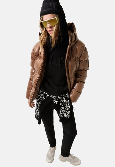 Person iført en brun dunjakke, sort hoodie, sort hue, store gule solbriller, mønstret sweater bundet om taljen, sorte bukser og hvide sneakers.