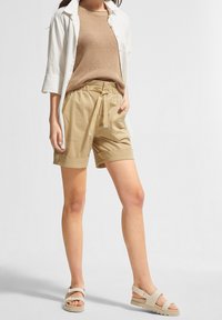 Beiget shortsit, joissa nyörireikä vyötäröllä ja taskut, yhdistettynä ruskeaan neulepuseroon ja valkoiseen nappipaitaan, täydentäen asua vaaleilla sandaaleilla.