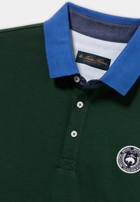 Dunkelgrünes Poloshirt mit blauem Kragen, versehen mit einem weißen Logo-Patch und Knopfverschluss. Glatter Stoff mit klassischem Design.