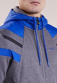 Felpa grigia con cappuccio imbottito blu, strisce nere e blu sulle spalle, chiusura a zip e dettaglio del logo sul petto. Materiali testurizzati utilizzati.