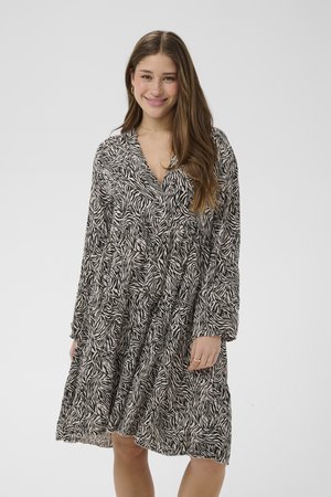 Jeune femme aux longs cheveux bruns portant une robe à imprimé zèbre noir et blanc, debout et souriante sur un fond clair uni.