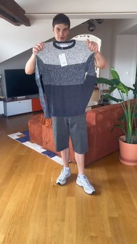 Muž stojí v obývacej izbe a drží šedý a námornícky modrý sveter s farebným blokom a cenovkou pred hrdzavohnedou pohovkou a rastlinou.