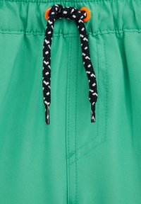 Tyrkysové krátke nohavice z látky s čierno-bielym pleteným šnúrkou prestrčenou cez oranžové očká na elastickom páse.