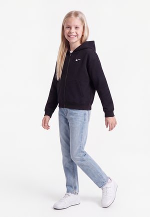 Fille souriante aux longs cheveux blonds, vêtue d'un sweat à capuche noir Nike, d'un jean bleu clair et de baskets blanches, debout sur un pied.