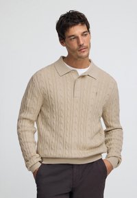 Beżowy sweter z dzianiny z wzorem warkocza, kołnierzykiem polo i ściągaczami na rękawach. Model nosi ciemne spodnie, utrzymując neutralną paletę kolorów.