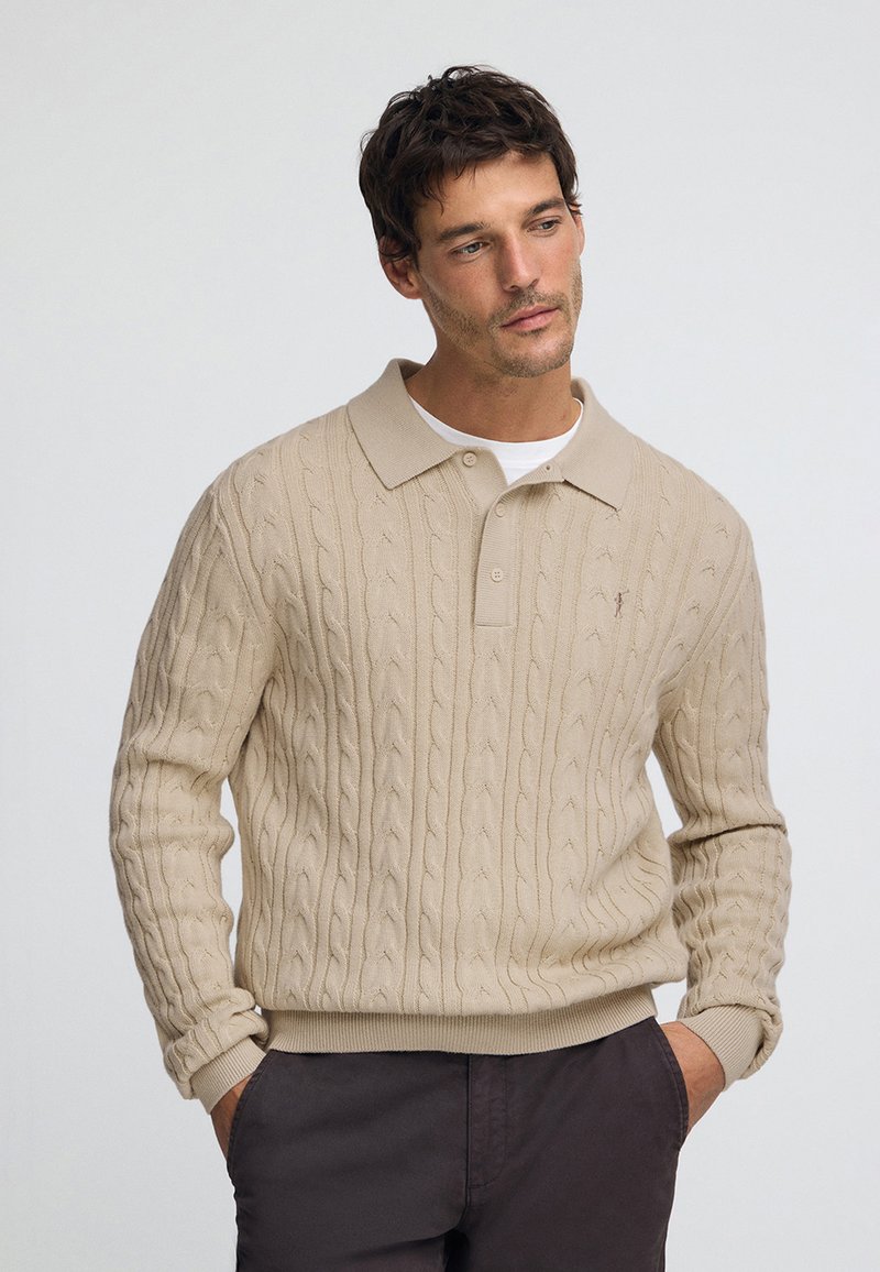 Beżowy sweter z dzianiny z wzorem warkocza, kołnierzykiem polo i ściągaczami na rękawach. Model nosi ciemne spodnie, utrzymując neutralną paletę kolorów.